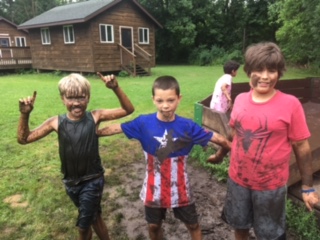 Three very muddy campers pose for a photo in front of a cabin