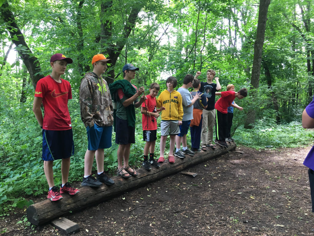 A line of boys of varied ages stands on a log in a wooded area.