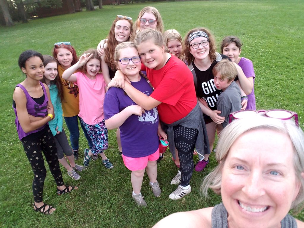 A woman takes a selfie with a large group of young girls at camp, many of them laughing and hugging.