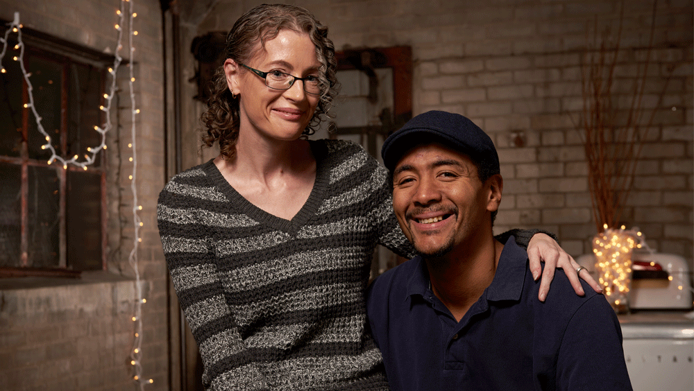 A woman sits on a man's lap in a festive room with exposed brick; both smile at the camera