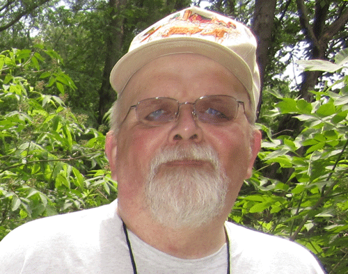 A man with a white beard and a baseball camp poses for the camera in front of camp greenery