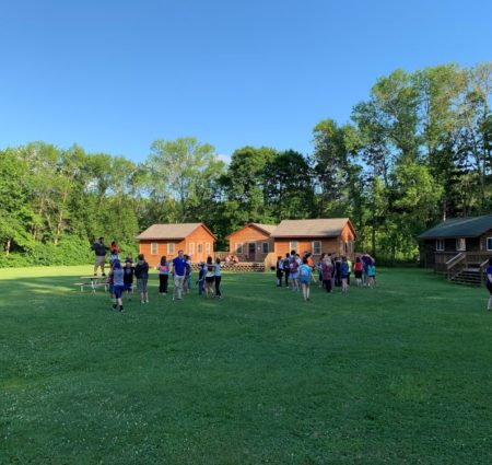 A large group of campers gather in a field in front of cabins at Camp Oz