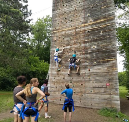 Several campers scale a climbing wall in harnesses while other campers look on