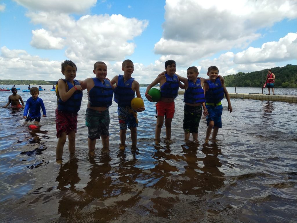 A group of campers in life preservers stand in ankle deep water